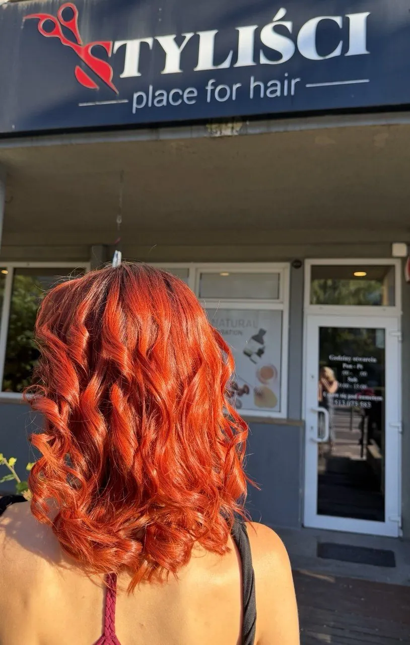 Miedziane loki klientki widziane od tyłu przed szyldem salonu Styliści na Marcelińskiej 54